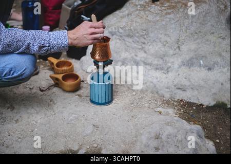 Un individuo gestisce una stufa a gas portatile, preparando caffè in una pentola di rame durante un viaggio in campeggio. Le tazze di legno riposano nelle vicinanze su terreni rocciosi. Foto Stock