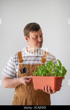 Il giardiniere che indossa una tuta marrone tiene una pentola di terracotta contenente piantine di basilico, pronta a piantarle nel suo giardino di casa, promuovendo una vita sostenibile e sana Foto Stock