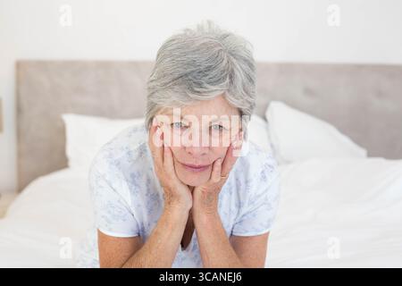 Donna anziana che riposa il mento sulle mani e guarda in avanti su un letto ben fatto con lenzuola bianche Foto Stock