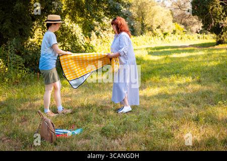 Felice famiglia caucasica che prepara una coperta da picnic sul prato nel parco soleggiato. Mamma e figlia si godono l'estate e il vero riposo all'aperto. Colpo largo. Foto Stock