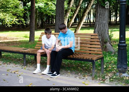 In un tranquillo parco, due giovani uomini sono seduti su una panchina di legno. Uno sta guardando un tablet, mentre l'altro guarda avanti. Sembrano essere dei reviewi Foto Stock