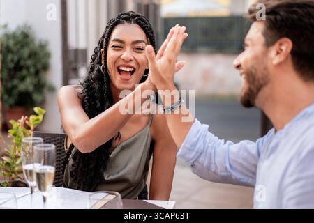 Una giovane coppia felice e variegata ride e regala cinque in festa mentre sorseggi vino frizzante in un caffè all'aperto. Un momento gioioso di successo, Foto Stock
