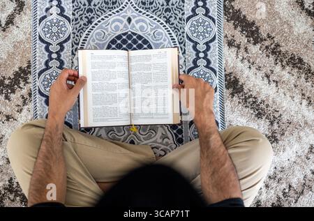Uomo musulmano che legge il Corano mentre siede su un tappeto di preghiera Foto Stock