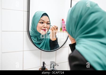 Donna in hijab che applica il trucco oculare nel bagno di casa Foto Stock
