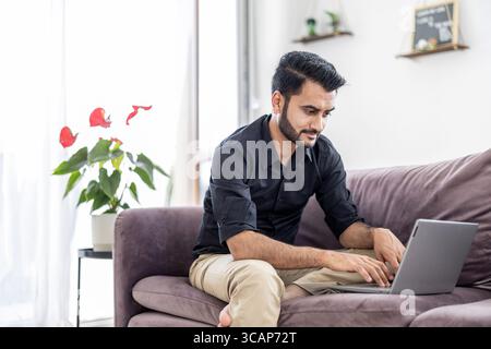 Uomo che utilizza un computer portatile seduto su un divano a casa Foto Stock