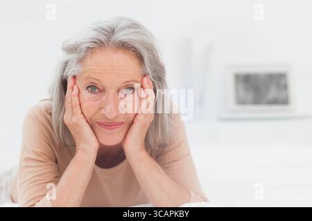 Donna anziana sdraiata sul letto in camera da letto, appoggiata alle mani tra i libri e concentrata sul laptop Foto Stock