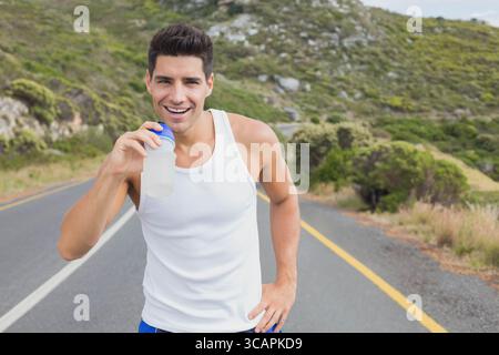 Corridore maschio in pausa su tortuosa strada asfaltata attraverso verdi colline, tenendo in mano una bottiglia d'acqua Foto Stock