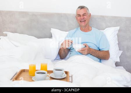 Un anziano seduto sul letto regge una ciotola in ceramica con cucchiaio accanto al vassoio della colazione e spazio per le copie Foto Stock
