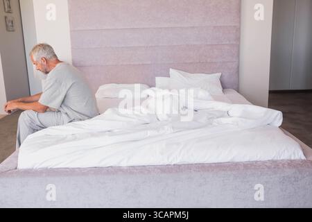 Uomo anziano seduto sul bordo di un letto non realizzato in una camera da letto dai toni pastello con testiera imbottita e spazio fotocopie Foto Stock