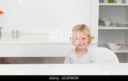 Bambino seduto a un tavolo in cucina tenendo la tazza accanto al vaso di fiori vicino agli scaffali, copia spazio Foto Stock