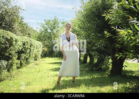 Un modello si erge graziosamente in un giardino verde circondato da alberi e cespugli vibranti sotto un cielo azzurro. Indossa un outfit elegante che mostra fash Foto Stock