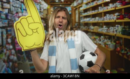 L'uomo tiene un dito in schiuma gialla e un pallone da calcio bianco e nero mentre tira in un colorato negozio di souvenir; fandom di entusiasmo. Foto Stock