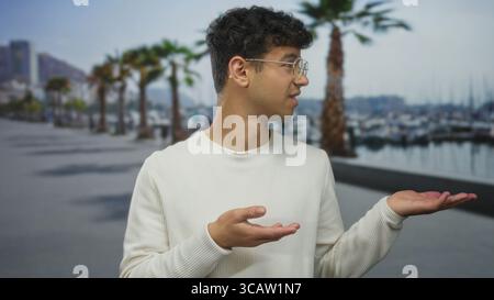 L'uomo che indossa un maglione bianco e occhiali mostra le palme rivolte verso l'alto accanto alla barca sul lungomare sotto le palme; incertezza. Foto Stock