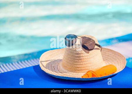 Concetto di vacanza estiva. Cappello di paglia con occhiali da sole e crema solare vicino alla piscina tropicale resort Foto Stock