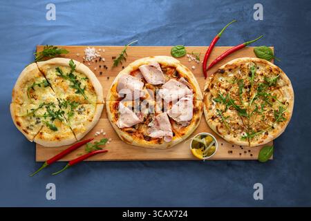 Una deliziosa esposizione di tre pizze su un vassoio di legno rustico, su un morbido sfondo blu, con condimenti vivaci e ingredienti freschi Foto Stock