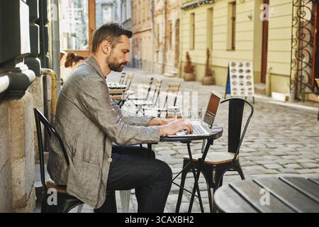 Uomo caucasico che lavora in remoto su un computer portatile in un bar all'aperto. Stile di vita freelance, concetto di nomade digitale Foto Stock