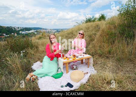 Due donne si godono un elegante picnic all'aperto con vino e spuntini, sorridendo su una collina estiva con vista panoramica sulla città. Foto Stock