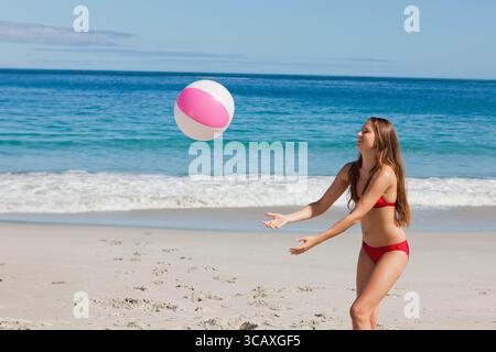 Donna che indossa un bikini rosso che lancia palla da spiaggia rosa sulla spiaggia di sabbia accanto al mare turchese, copia spazio Foto Stock