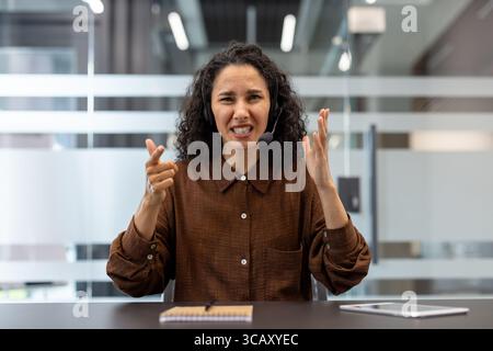 Una donna professionista mostra frustrazione durante una telefonata, seduta alla scrivania di un ufficio. I suoi gesti espressivi catturano le sfide delle interazioni con il supporto clienti e dello stress sul luogo di lavoro Foto Stock