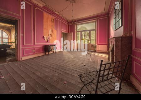 Camera rosa abbandonata con pareti sbucciate, mobili d'epoca e luce del sole che scorre attraverso grandi finestre in un vecchio castello Foto Stock