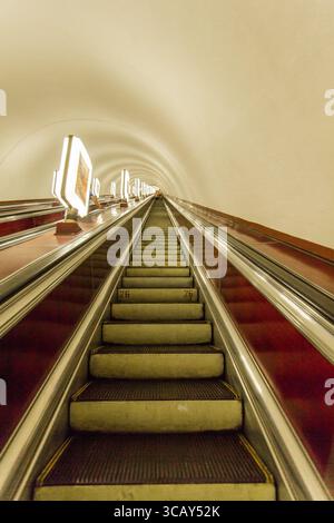 Lunga scala mobile all'interno di un tunnel con pubblicità illuminate e un design moderno, vista prospettica verso l'alto - metropolitana di Kiev e rifugio Ucraina Foto Stock