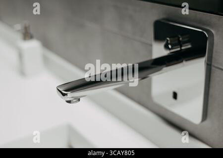 Un moderno rubinetto cromato in un bagno di lusso, con un sensore per l'uso senza contatto. Primo piano di un moderno rubinetto cromato montato su una parete piastrellata grigia, con una Foto Stock