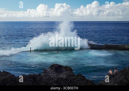 La potente onda oceanica si schianta in una piscina di roccia naturale in una giornata di sole, mentre le persone nuotano e si rilassano sulle rocce vulcaniche sotto un cielo blu con soffici nuvole Foto Stock