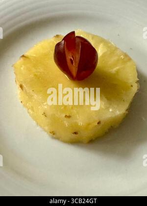 Fetta di ananas fresco condita con uva rossa tagliata a metà, servita su un piatto bianco. Foto Stock