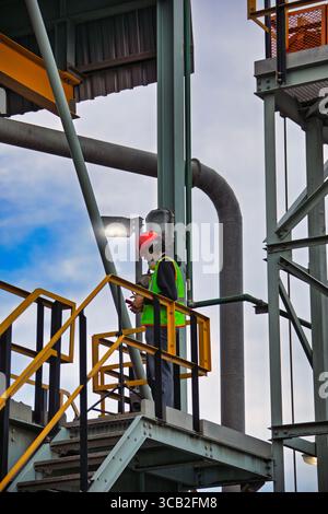 uomo appaltatore lavoratore ha scalato la struttura metallica industriale con attrezzatura di sicurezza e elmetto utilizzando il telefono, indossando indumenti da lavoro Foto Stock