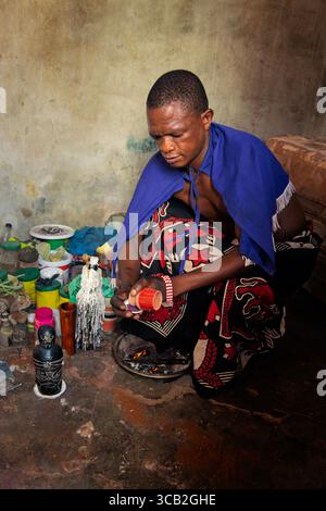 Sangoma del villaggio africano che indossa un mantello blu e un abbigliamento tradizionale, eseguendo un rituale spirituale all'interno, circondato da erbe, ossa e candele, B. Foto Stock