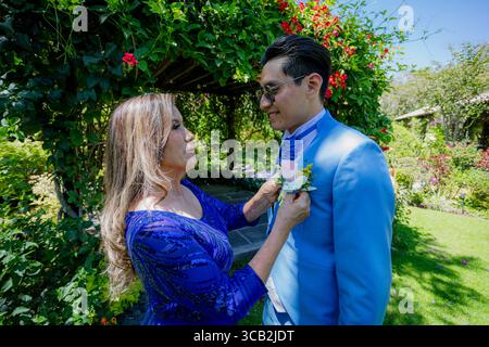 In un giardino soleggiato, la madre aiuta suo figlio con un abito blu ad adattare il fiore all'occhiello prima della cerimonia di nozze. Foto Stock