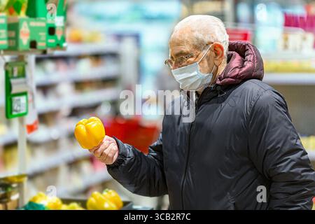 Vecchio europeo anziano che indossa una maschera protettiva per il viso che guarda il pepe paprika giallo al supermercato. Shopping durante il concetto di COVID-19 Foto Stock