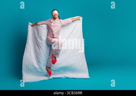 Bella donna che posa allegramente in pigiama rosa con piumone e maschera dormiente su uno sfondo blu brillante Foto Stock