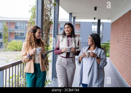 Diversi colleghi che indossano abiti da lavoro che camminano lungo il passaggio dell'ufficio all'aperto con tazze da caffè Foto Stock