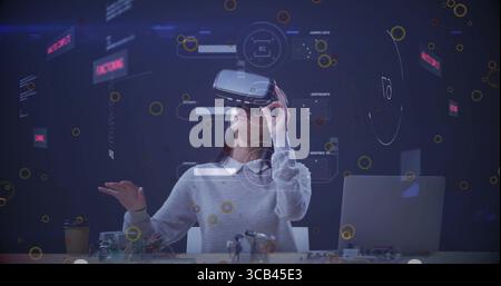 Tecnico addetto ai test che indossa il visore VR alla scrivania del laboratorio, con laptop, tazza da caffè, elettronica e ologrammi Foto Stock