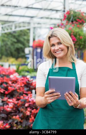 Lavoratrice vivaistica che indossa grembiule verde che tiene in mano la compressa mentre ispeziona i fiori rossi in serra Foto Stock