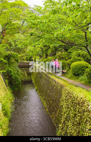 Due persone si godranno una tranquilla passeggiata lungo il sentiero del filosofo lungo il pittoresco canale circondato da vibranti alberi verdi, Kyoto, Giappone Foto Stock