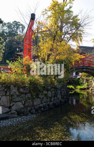 Vivace scena di un sereno giardino del Santuario di Shimogamo caratterizzato da una porta torii rossa, un ponte in pietra e lussureggiante fogliame autunnale lungo un limpido ruscello sotto il ruscello Foto Stock