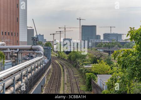 Binari ferroviari con edifici industriali e numerose gru da costruzione sullo sfondo. Area di sviluppo urbano con architettura moderna e verde Foto Stock