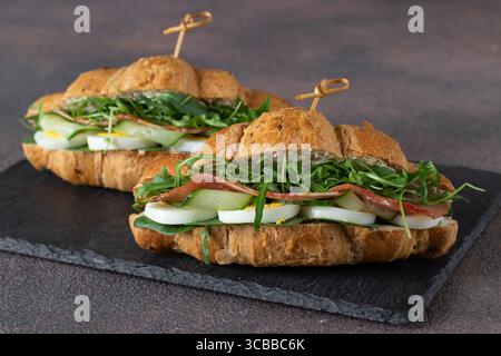 Panini con croissant con prosciutto, cetriolo, uovo e rucola su piatto di ardesia Foto Stock