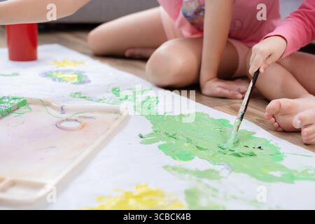 Due sorelle sedute sul pavimento del soggiorno, dipingono insieme su un grande foglio di carta con pitture e pennelli colorati. Foto Stock