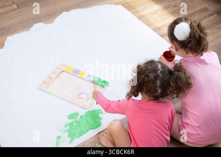 Due sorelle sedute sul pavimento del soggiorno, dipingono insieme su un grande foglio di carta con pitture e pennelli colorati. Foto Stock