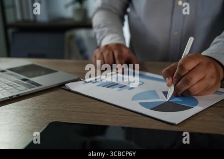 Primo piano di un imprenditore online che analizza grafici di dati su carta con penna stilo accanto al notebook, che rappresenta la strategia aziendale digitale, il lavoro remoto, un Foto Stock