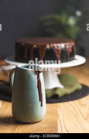 Una caraffa in ceramica con salsa di cioccolato che gocciola lateralmente, con una cheesecake ricoperta di ganache di cioccolato lucido sullo sfondo. Foto Stock