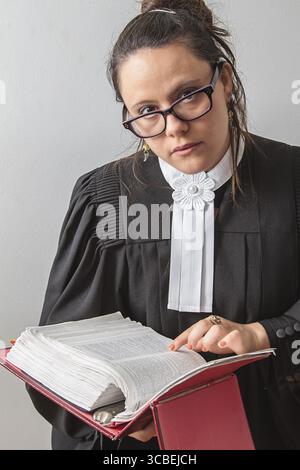 Trenta qualcosa di bruna donna che indossa una toga canadese da avvocato con un libro rosso di diritto penale in mano che guarda i suoi occhiali Foto Stock