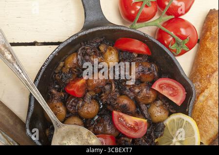 Baby seppie arrostite in padella di ferro con pomodori e cipolle su tavola in legno rustico Foto Stock
