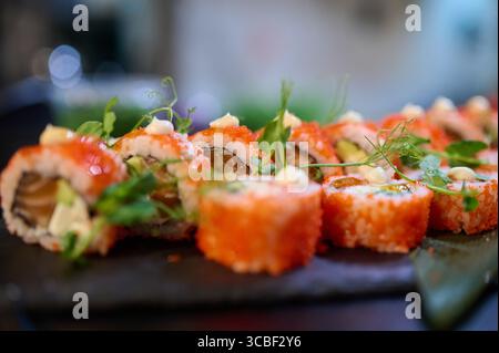 Primo piano di deliziosi panini di sushi conditi con tobiko, salsa di panna e microgreen, disposti su un piatto di ardesia nera in un ristorante giapponese Foto Stock