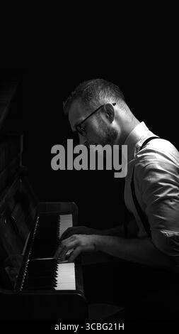 Uomo che suona il pianoforte con un'illuminazione soffusa e scura con occhiali e bretelle Foto Stock