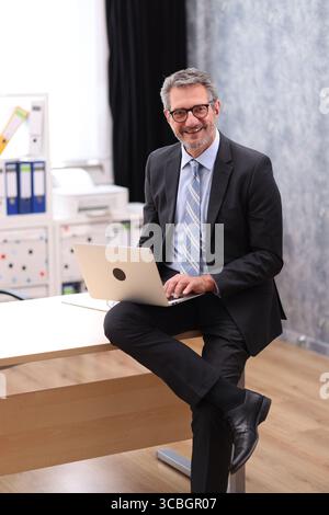 Uomo d'affari in tuta nera seduto in ufficio e che lavora su un computer portatile Foto Stock
