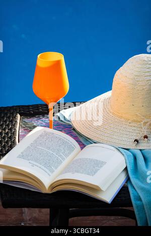 Un perfetto scenario di vacanza estiva con un libro aperto, un cappello di paglia e un drink colorato seduto su un tavolo accanto a una piscina blu brillante in un lussuoso hotel Foto Stock